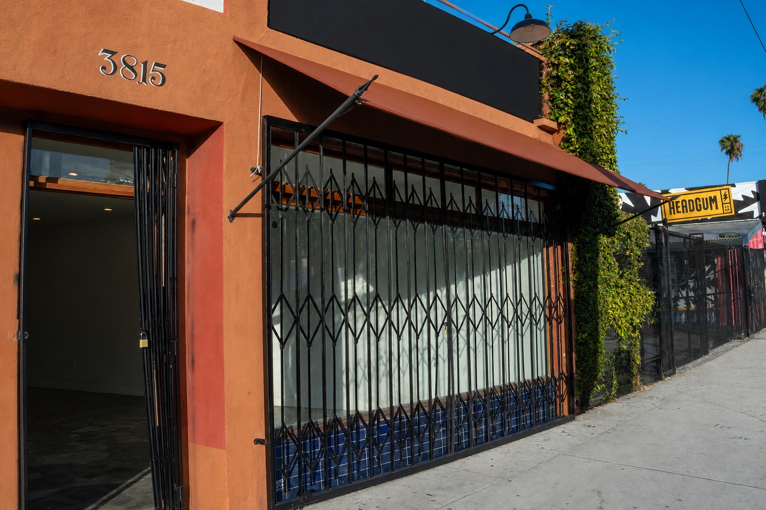 The available 3815 W. Sunset Blvd. storefront in its current condition, seen from the sidewalk: terracotta façade, folding security gate across the display window, open entry door, and the painted street number 3815.