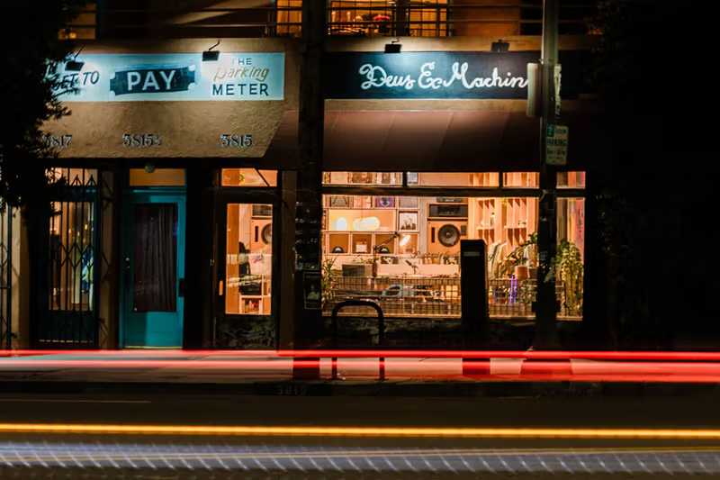 The 3815 W. Sunset Blvd. storefront during its prior occupancy by Deus Ex Machina, lit at night with the tenant's signage and window display in place.