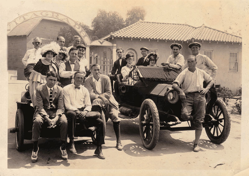 Archival black-and-white photograph of a group of people posed with two early automobiles beneath an arch lettered Vitagraph, in front of a tile-roofed building.