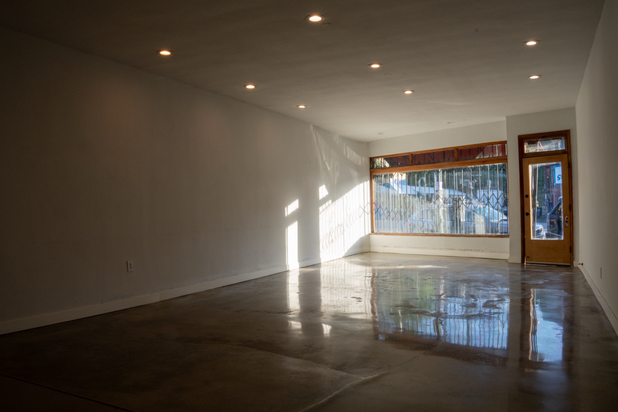 Daylight entering the vacant 3815 W. Sunset Blvd. interior through the full-width storefront window and timber entry door, reflected on the polished concrete floor.