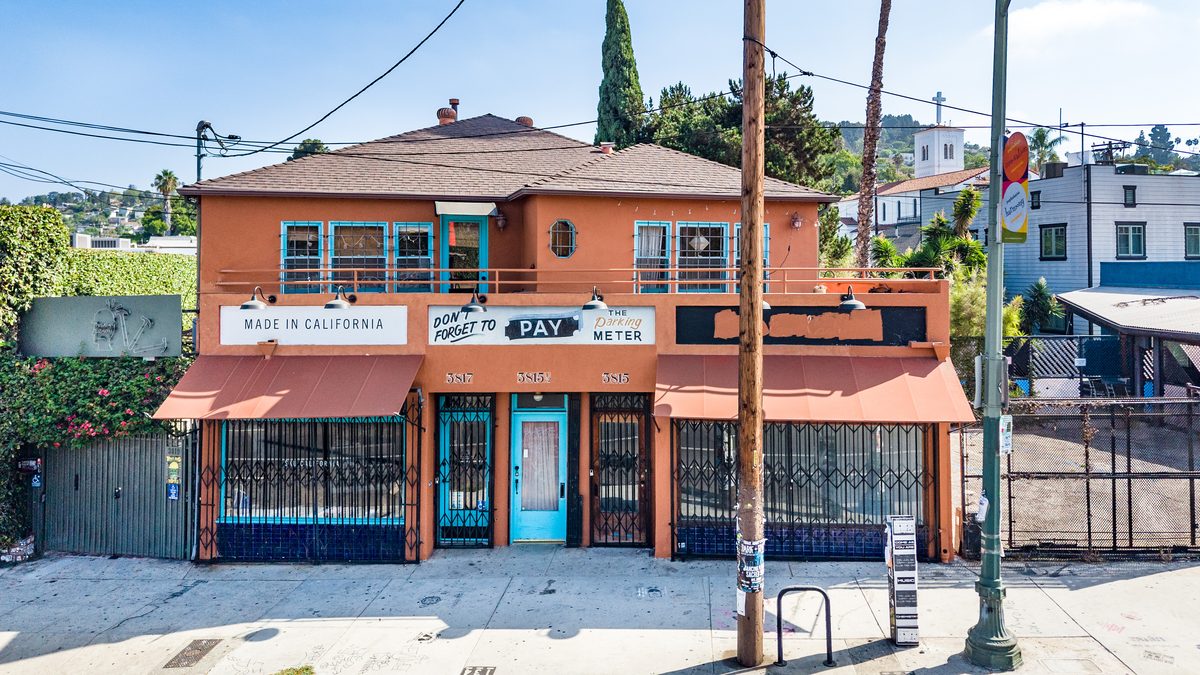 The two-storey terracotta building at 3815–3817 W. Sunset Blvd., seen from across the sidewalk: red-tile awning, steel security gates, a pale-blue center door, and painted street numbers 3817, 3815½, and 3815. The available suite is the right-hand storefront at 3815.