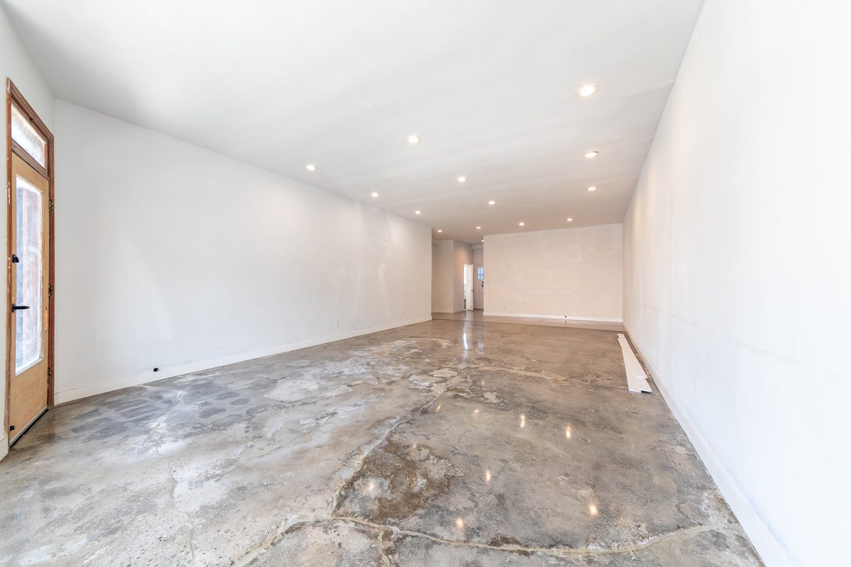 Vacant main room of 3815 looking in from the storefront: polished concrete, white walls, recessed ceiling lights, timber door at left, and a rear opening at the far wall.
