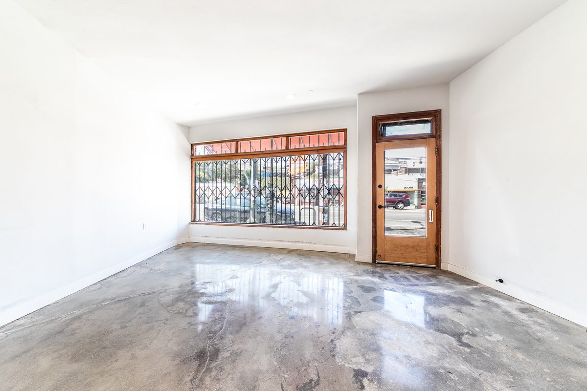 Vacant main room looking toward the storefront: gated display window, timber entry door with a transom, white walls, and polished concrete.