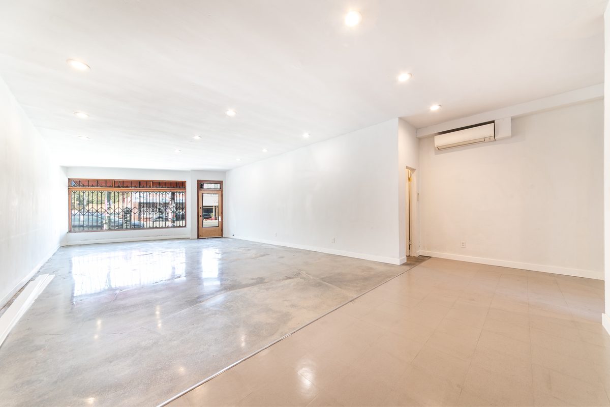 Wide view of the vacant main room: storefront window and timber door at left, white walls, polished concrete, recessed lights, and a wall-mounted air conditioner.