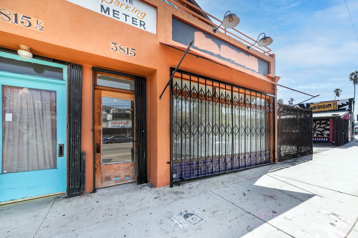Close sidewalk view of the 3815 entry: painted street number 3815, timber door with a kick plate, and a folding security gate across the display window.